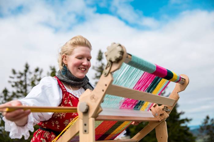 Onnellisen näköinen nainen kutomassa värikästä huivia matkakangaspuilla kauniissa Koli-maisemassa. 